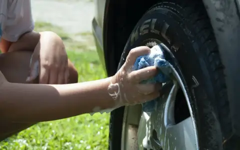 A person's arm and hand, covered in suds, scrub a car tire with a blue sponge. It seems they are testing if you can use dish soap to wash your car.