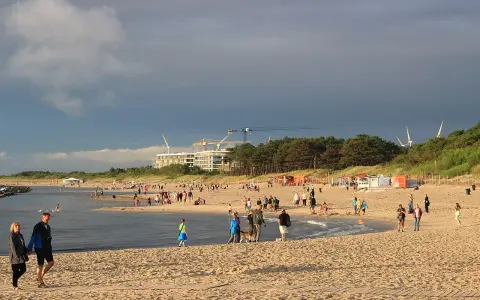 Nadmorskie miasto nad Wieprzą tętni życiem. Ludzie spacerują po plaży, bawią się w wodzie, a w tle widać budowę i wiatraki.