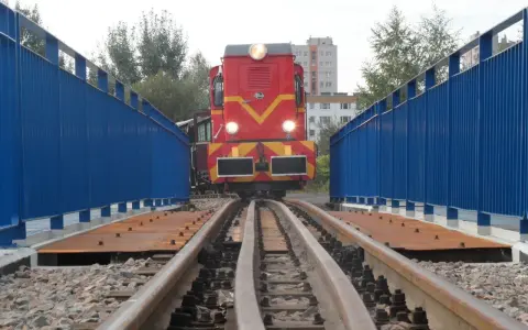 Czerwona lokomotywa na torach wąskotorowych, część górnośląskich kolei wąskotorowych, wjeżdża na most z niebieskimi barierkami.