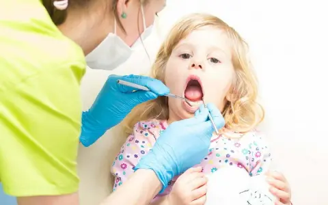 Mała dziewczynka z otwartymi ustami, kiedy z dzieckiem do dentysty. Stomatolog w niebieskich rękawiczkach bada jej ząbki.