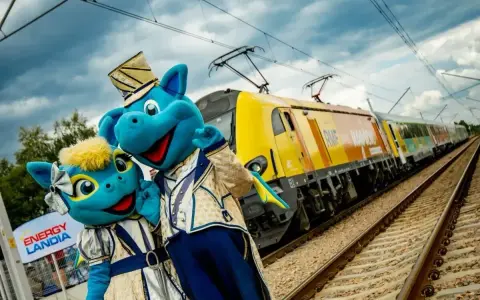 Dwa niebieskie smoki w strojach stoją obok żółtego pociągu na torach kolejowych.