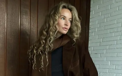 Kobieta z kręconymi, beżowymi włosami, którym pasuje ten odcień. Elegancka stylizacja z brązową marynarką.