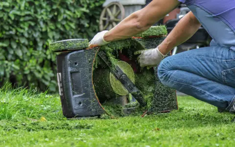 Osoba w białych rękawiczkach dokonuje wymiany noża w kosiarce, która jest pochylona na trawie.