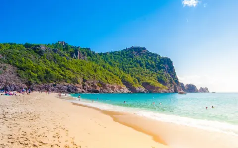 Plaża z turkusową wodą, zielonymi wzgórzami i ruinami zamku na szczycie.
