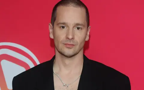 Krzysztof Zalewski w czarnym garniturze, z naszyjnikiem, na tle czerwonym z białym logo.
