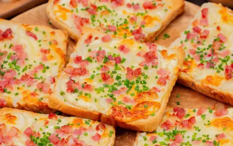 Mehrere Flammkuchen-Toast-Scheiben mit Speck, Zwiebeln und Schnittlauch belegt, auf einem Holzbrett.