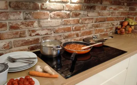 Cegła w kuchni inspiracje: gotowanie na płycie indukcyjnej, obok garnki, patelnia z sosem, warzywa i owoce.