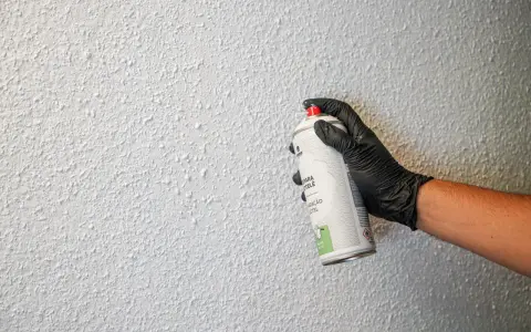 Mano con guante negro aplica spray para reparar gotelé. La pared tiene textura rugosa y blanca.