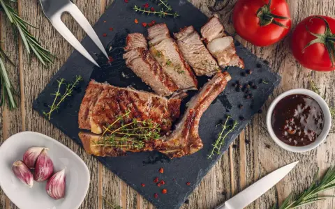 Côte de veau grillée tranchée sur une planche d'ardoise, garnie d'herbes, avec des tomates, de l'ail et une sauce.