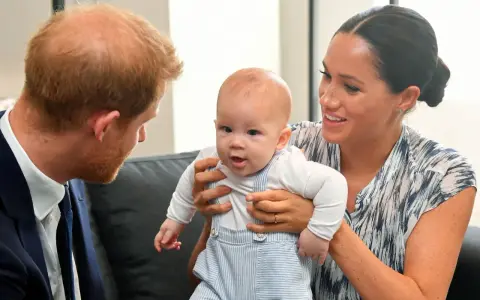 Meghan Markle håller sitt barn, som tittar nyfiket framåt. Prins Harry ser på dem med ett leende.