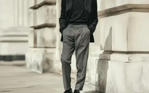 Uomo con outfit elegante: giacca scura, maglione nero e pantaloni chino grigi. Scarpe stringate lucide completano il look.