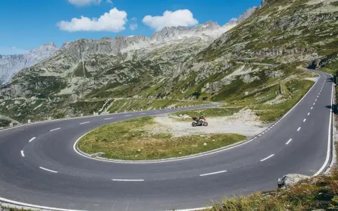 Una moto attende su un tornante del Grimsel Pass, tra montagne verdi e cielo azzurro.