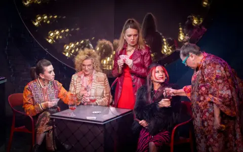 Bożena Zawiślak Dolny w czerwonej sukience i skórzanej kurtce trzyma karty. Obok siedzą kobiety przy stole z drinkami.