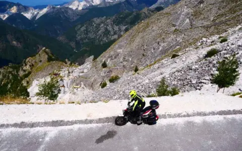 Motociclista in abbigliamento giallo fluo affronta il passo del vestito, tra cave di marmo e montagne imponenti.