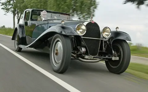Ein Mann fährt einen glänzenden schwarzen Bugatti Royale auf einer Landstraße. Der Bugatti Royale Preis ist astronomisch, ein Traum für jeden Sammler.