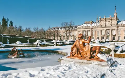 Schneebedeckte Gärten und ein gefrorener Brunnen vor einem Palast in Spanien im Winter. Eine bronzene Statue blickt auf die verschneite Landschaft.