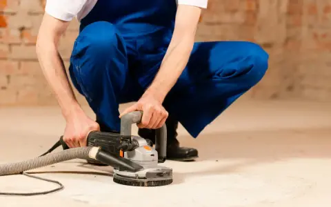 Mann in blauer Latzhose schleift mit einer Maschine den Estrich. Staub wird abgesaugt.