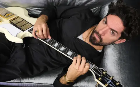 El cantante Siloe, con barba y cabello oscuro, toca una guitarra eléctrica blanca con detalles dorados, recostado en un sofá de terciopelo gris.