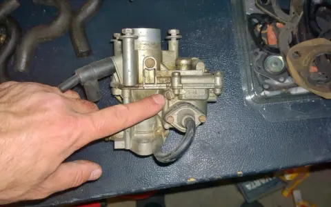 Main d'homme pointant un carburateur Zenith, possible cause d'un moteur 4 temps qui s'étouffe à l'accélération. Pièces de rechange visibles.