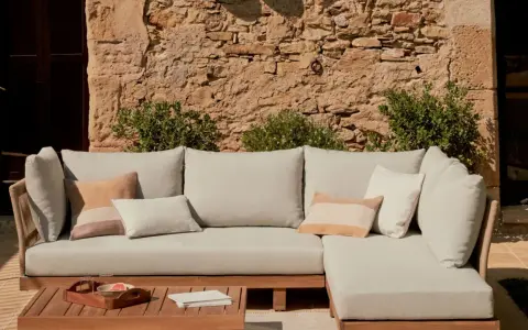 Sofá modular de exterior con cojines en tonos neutros, sobre una mesa de madera. La **decoración estilo natural** se realza con la pared de piedra y la vegetación.