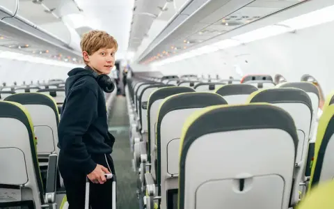 Chłopiec z słuchawkami na szyi i walizką w ręku stoi w przejściu samolotu. Od ilu lat można latać samemu? Ryanair pozwala na samodzielne podróże od 12 roku życia.