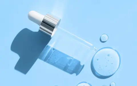 En pipett med klar vätska, som innehåller hyaluronsyra och retinol, ligger på en ljusblå bakgrund med droppar av serum.