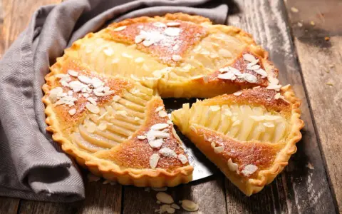 Une délicieuse tarte aux poires, garnie d'amandes effilées et saupoudrée de sucre glace, repose sur un plateau en bois.