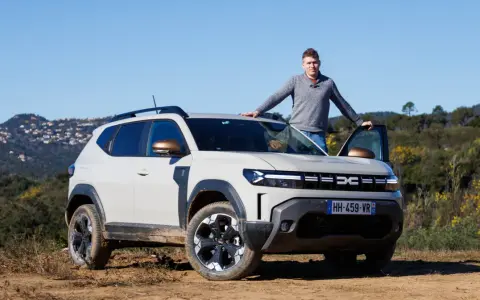 Mężczyzna opiera się o maskę jasnoszarego Dacia Duster, hybrydy z fabrycznym LPG, na tle wzgórz.