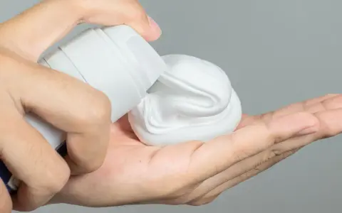Mano aplicando gel o espuma de afeitar de un envase blanco a la palma.