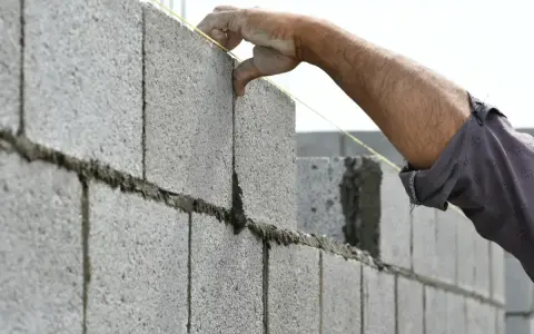 Main d'un maçon plaçant une brique sur un mur en parpaing, guidé par un fil à plomb.
