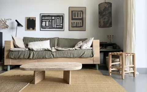 Une idée table basse fait maison en bois brut, posée sur un tapis tressé, devant un canapé vert et des tableaux abstraits.