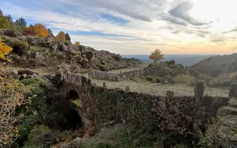 Herbstliche Farben in der Sierra de Gata. Ein alter Steinbogenbrücke, überwuchert von Efeu, führt durch eine felsige Landschaft.