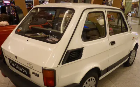 Biały Fiat 126p bis z naklejkami na tylnej szybie, stojący obok czerwonego samochodu.