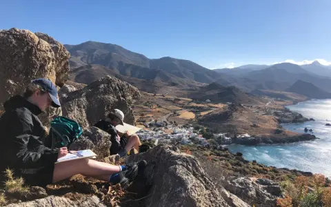 Två personer sitter på en klippa och njuter av utsikten över en by vid havet, inspirerade att måla landskap.