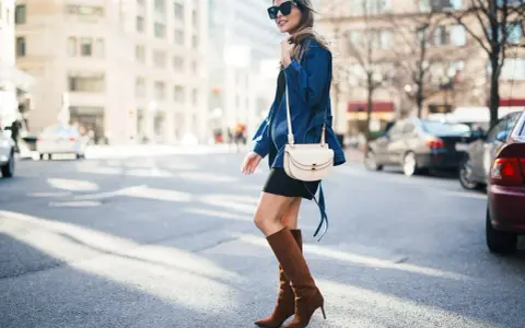 A woman walks down a city street wearing a black dress, a denim jacket, and stylish brown boots.