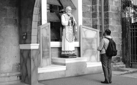Pomnik Ojca Pio, patrona modlących się o uzdrowienie, stoi przed kościołem. Mężczyzna z plecakiem patrzy na tablicę.