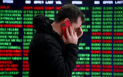Hombre preocupado habla por teléfono frente a pantalla de bolsa de valores de Nueva York, con números rojos y verdes indicando un posible crack.