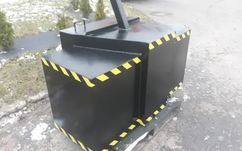 Czarny, metalowy balast do ciągnika z żółto-czarnymi pasami ostrzegawczymi, stojący na drewnianej palecie.