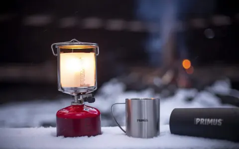 En varm primus lampa lyser upp snön bredvid en mugg och en termos. I bakgrunden syns en lägereld.