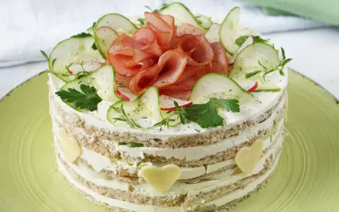 Herzhafter Kuchen mit Schichten aus Brot, Frischkäse, Gurken und Schinken. Dekoriert mit Radieschen, Kräutern und Käseherzen.