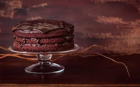 Torta al cioccolato a tre strati, lucida e invitante, su un'alzata di vetro. Gocce di glassa scivolano sui bordi.