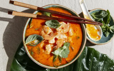 Miseczka zupy tom yum z krewetkami, ozdobiona ziołami, pałeczki i łyżka.