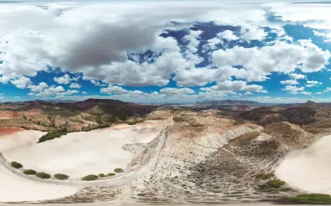 Paisaje árido con colinas, campos cultivados y un camino serpenteante. El cielo azul con nubes blancas contrasta con el desierto de larva.