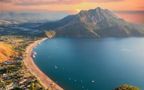 Malownicze wybrzeże Turcji o zachodzie słońca. Góry schodzą do turkusowego morza, a plaża z leżakami i łodziami zaprasza do odpoczynku.