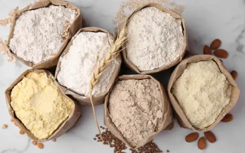 Six sacs en papier remplis de différentes farines, dont une farine de blé CRC. Amandes et grains parsèment le fond marbré.