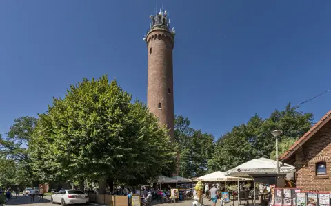 Wysoka, ceglana latarnia morska Sarbinowo góruje nad letnim kurortem z drzewami i ogródkami kawiarnianymi.