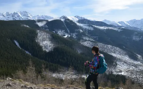 Widok na wielki kopieniec górski pokryty lasem i śniegiem. Kobieta z plecakiem i kijkami trekkingowymi podziwia panoramę.