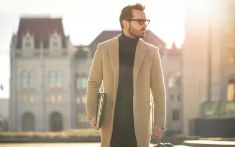 Ein Mann im old money style, mit Brille und beigem Mantel, trägt einen Laptop und eine Reisetasche.