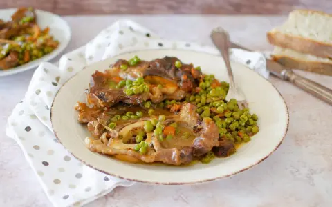 Un piatto di ossobuco con piselli e carote, servito su un piatto bianco con una forchetta. Sullo sfondo, fette di pane.