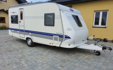 Biała przyczepa kempingowa Hobby 460 UFE "de luxe easy" stoi na kostce brukowej przed żółtym budynkiem.